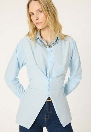 Femme portant une chemise bleu clair boutonnée avec des pans avant prolongés et un jean bleu foncé, accessoirisée d’un collier argenté conspicueux.