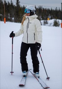 Skioutfit: witte waterdichte jas met zwarte accenten, zwarte skibroek, zwarte handschoenen, blauwe gemirroreerde goggles en witte helm, op een besneeuwde helling.