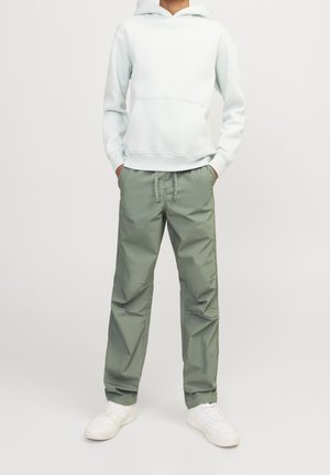 Personne portant un sweat à capuche blanc, un pantalon à cordon de serrage gris-vert clair et des baskets blanches, se tenant devant un fond clair uni.