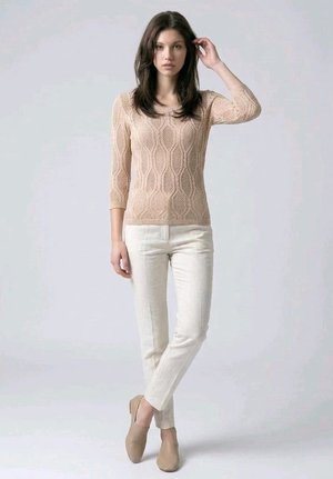 Mujer de pie con una mano tocándose el pelo, vistiendo un suéter de punto beige, pantalones blancos y zapatos beige sin cordones, sobre un fondo liso.