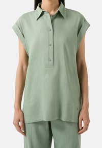 Donna che indossa una blusa verde salvia a maniche corte e sfonda larga, con colletto e pantaloni abbinati, in piedi di fronte a uno sfondo neutro.