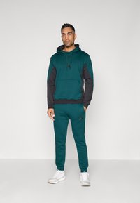 Zeleno-modra kapuca z črnimi detajli in sprednjim žepom, ujemajoča se z zeleno-modrimi trenirkami s logotipom Adidas, izdelana iz mehke, teksturirane tkanine.