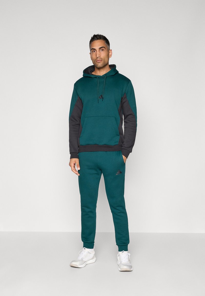 Zeleno-modra kapuca z črnimi detajli in sprednjim žepom, ujemajoča se z zeleno-modrimi trenirkami s logotipom Adidas, izdelana iz mehke, teksturirane tkanine.