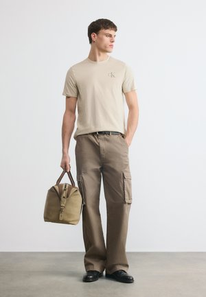 Άνδρας που φοράει μπεζ μπλουζάκι και καφέ παντελόνι cargo κρατά μια χακί σακ βουαγιάζ, στέκεται μπροστά σε ένα απλό φωτεινό φόντο.