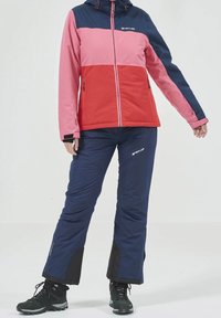 Giacca invernale a blocchi di colore con pannelli rosa, rossi e blu navy, abbinata a pantaloni isolati blu navy e stivali da trekking neri con accenti grigi.