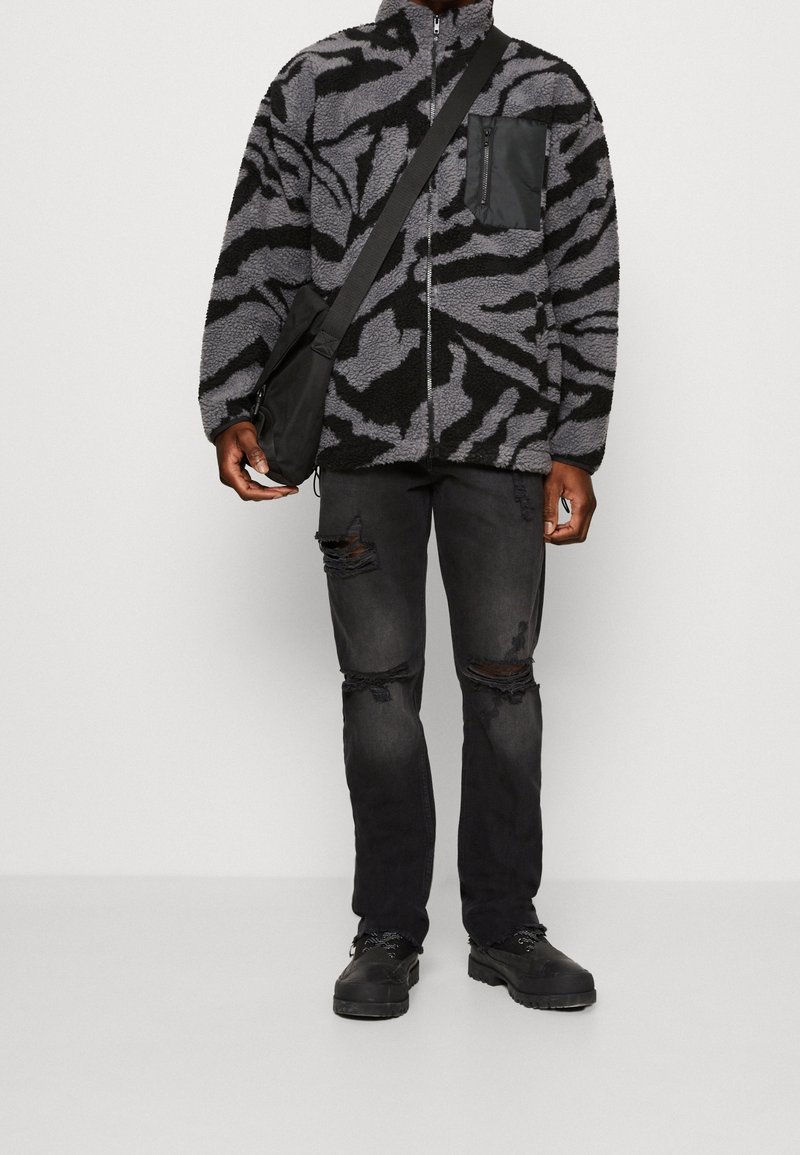 Personne portant une veste polaire à motif zèbre noir et gris, un jean noir déchiré, des chaussures noires, et portant un sac noir en bandoulière.