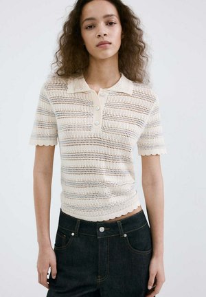 Joven mujer con un polo de punto crema de manga corta con bordes ondulados y vaqueros oscuros de cintura alta sobre un fondo liso.