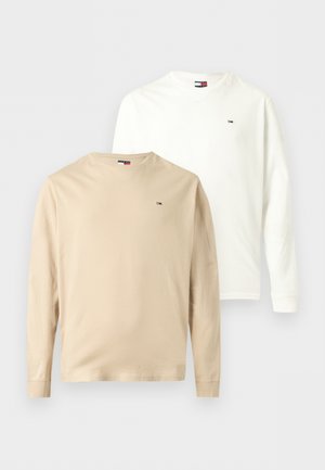 Tânăr cu păr închis la culoare, purtând o cămașă albă cu mâneci lungi, blugi largi bleu deschis și pantofi maro închis, stând cu mâinile în buzunare.