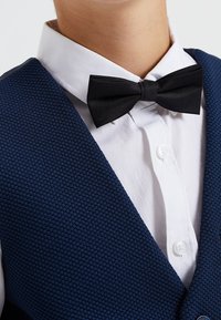 Marinblå texturerad väst med vit skjorta under, utrustad med en svart sidenfluga och en knappdetalj på västens högra sida.