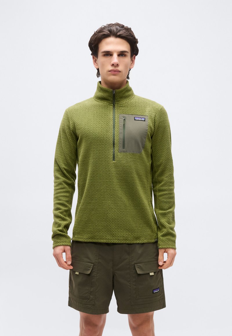 Νέος άνδρας που φοράει φλις ελιάς με υφή και φερμουάρ μέχρι το στήθος, με τσέπη στο στήθος και σκούρα πράσινα cargo σορτς, στέκεται μπροστά σε απλό φόντο.