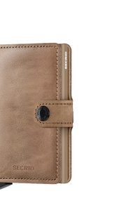 Cartera de cuero marrón con superficie texturizada, bordes cosidos, cierre de botón en la parte superior y un detalle de marca en el lateral. Diseño rectangular compacto.