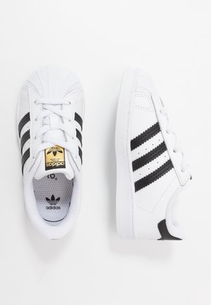 Par hvide Adidas-sneakers med sorte striber, shell toe-design og gyldent logo på tungen, vist fra top- og sidevisning.