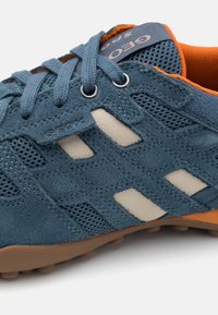 Baskets en daim bleu avec panneaux en maille, accents beiges, doublure intérieure orange, et semelle en caoutchouc texturée. Comprend des lacets ronds et une étiquette logo.