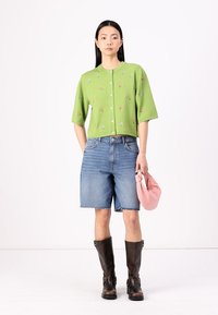 Grønn kortermet cardigan med broderte blomsterdetaljer, kombinert med denim bermudashorts. Mørkebrune knelange støvler og en rosa veske.