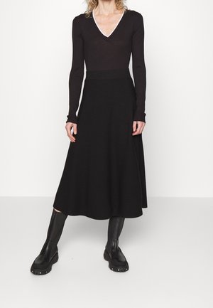 Woman wearing black long-sleeve V-neck top, black midi skirt, and black knee-high chunky boots, standing against a plain white background.