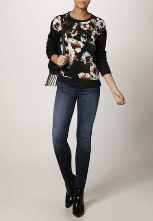 Vrouw die een zwarte sweatshirt met bloemenprint draagt, donkerblauwe skinny jeans, zwarte enkellaarsjes en een zwarte schoudertas met franjes.