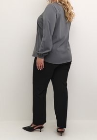 Kvinde med langt blond hår iført en grå bluse med lange ærmer, sorte straight-leg bukser og sorte spidse kitten heels.