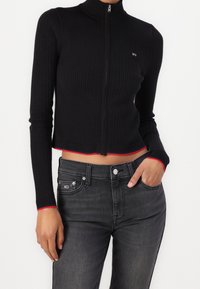 Černý žebrovaný svetr na zip s vysokým límcem. Má červené akcenty na manžetách a spodním lemu. Kombinován s tmavě šedými džíny.