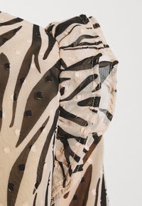 Texturiertes Gewebe mit einem beigefarbenen und schwarzen Zebra-Muster, das ein Rüschendetail an der Schulter und kleine bestickte Punkte überall aufweist.