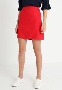 Jupe rouge longueur genou en tissu lisse avec une taille ajustée et un devant plat. Assortie à des chaussures blanches sans lacets, offrant un look décontracté.