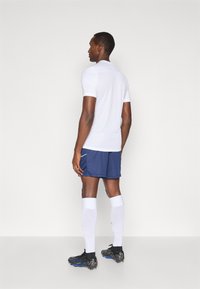 Αθλητική ενδυμασία που χαρακτηρίζεται από ένα λευκό κοντομάνικο polo, ναυτικές βερμούδες, λευκές κάλτσες μέχρι το γόνατο και μαύρα αθλητικά παπούτσια με μπλε λεπτομέρειες.