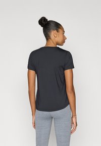 Μαύρο κοντομάνικο T-shirt με στρογγυλή λαιμόκοψη, κατασκευασμένο από ομαλή υφασμάτινη ύλη, φορεμένο με ανοιχτό γκρι κολάν. Η οπίσθια όψη δείχνει καμπυλωτή γραμμή τελειώματος.