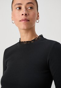 Black ribbed long-sleeve top with a textured black lace ruffled neckline. Features a fitted silhouette and smooth fabric texture.