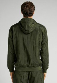 Chaqueta ligera de color verde oliva con una textura arrugada, capucha y puños y bajo acanalados, con un diseño minimalista y sin gráficos visibles.