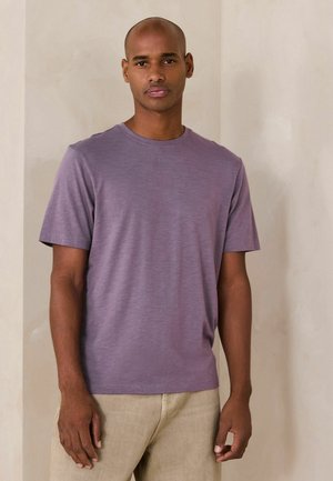 Vīrietis ar neitrālu izteiksmi, valkājot vienkāršu violeta īsroku krekli un bēšas bikses, stāvot pret gaišu sienas fona.