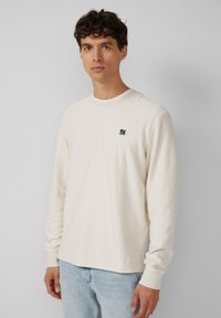 Crème langemouw T-shirt van textuurstof, ronde halslijn en een klein marineblauw logo op de linkerborst, gecombineerd met lichtblauwe spijkerbroek.
