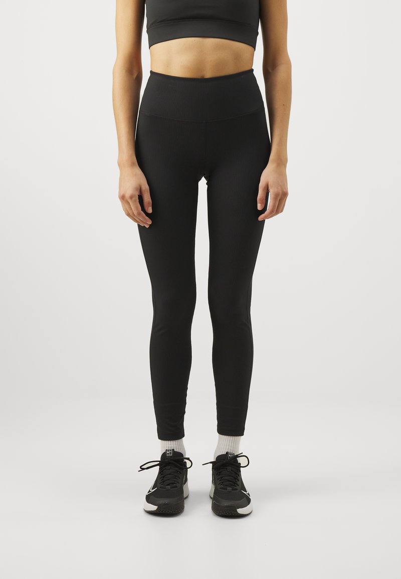 Svarta leggings med hög midja, gjorda av ett ribbat tyg, kombinerade med svarta träningsskor och vita strumpor. Slät textur, figurnära design.