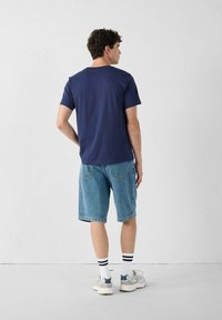 Navyblauw katoenen t-shirt met korte mouwen, gecombineerd met lichtblauwe denim shorts. Model draagt witte sokken met zwarte strepen en grijze sneakers.