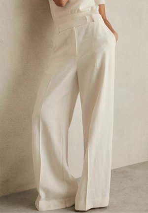 Mujer con pantalones blancos de talle alto y pata ancha con bolsillos laterales, de pie contra una pared y suelo color beige claro.