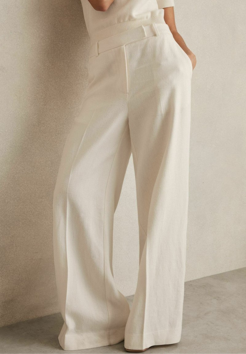 Femme portant un pantalon blanc taille haute à jambes larges avec poches latérales, debout contre un mur et un sol beige clair.