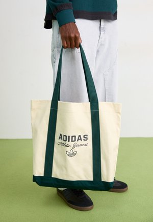Person holding beige Adidas tote bag with dark green handles, wearing light gray pants and black sneakers on green floor.