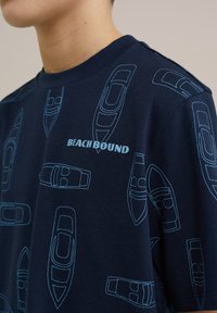 Tamsiai mėlyna marškinė blizgusiai šviesiai mėlynos valties grafiką su užrašu "KRYPSTAME Į PAPLAUDIMĮ." Pagaminta iš minkštos medvilnės su klasikiniu apvaliu iškirpimu.