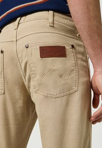 Beżowe dżinsy Wrangler z brązową skórzaną metką na tylnej kieszeni, z kontrastowym przeszyciem i kieszenią w kształcie diamentu.