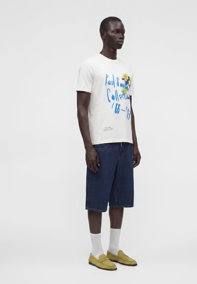 Άνδρας που φοράει άσπρο γραφικό t-shirt, σκούρα μπλε βερμούδα μέχρι το γόνατο, λευκές κάλτσες και κίτρινα μοκασίνια στέκεται μπροστά σε απλό φόντο.