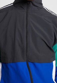 Färgglad zip-up jacka med svart överdel, teal ärmar och blå nedre del med vita accenter och tre vita ränder på axlarna.