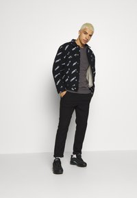 Svart jakke med hvit «OBEY» trykk, grå t-skjorte, svarte bukser og svarte sneakers. Modellen står med hendene i lommer, kort lyst hår.
