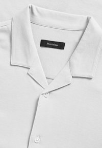 Lichtgrijs polo shirt met een zachte textuur, voorzien van een puntige kraag, twee witte knopen en een zwart label met de tekst "Matinique."