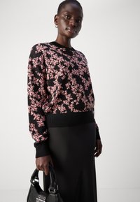 Črna pulover s roza cvetličnim vzorcem, rebrastimi manšetami in robom, v kombinaciji s črno krilo in v roki drži črno torbico. Gladka tekstura.