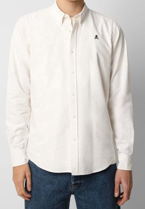 Homme portant une chemise blanche boutonnée avec un logo tête de mort et os croisés noirs sur la poitrine et un jean bleu.