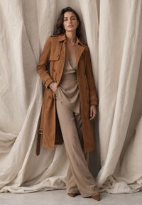 Frau steht vor beige drapiertem Stoff, trägt einen braunen Wildleder-Trenchcoat über einem beigen Wickeltop und weit geschnittenen Hosen.