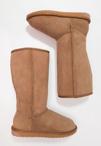 Par de botas de invierno marrones de ante hasta la mitad de la pantorrilla con costuras visibles y suelas de goma, una bota en posición vertical y la otra tumbada de lado.