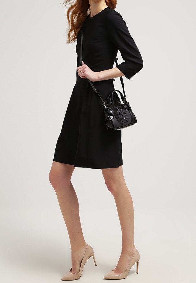Femme portant une robe noire longueur genou, des talons hauts beiges, et un petit sac à main noir avec une bandoulière.