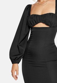 Černé šaty s přiléhavým siluetou, nabíranými dlouhými rukávy a stahovaným vrškem s odhalenou střední částí.