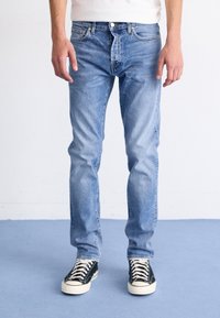 Jeans de denim azul claro con un ajuste slim, diseño de cinco bolsillos, costuras clásicas y cierre de botón. Combinados con zapatillas negras y blancas.