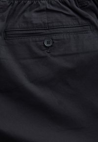 Juodos medžiagos su lygiu paviršiumi skyla mygtuko užsegimu virš nugaros kišenės. Siūlės matomos palei kraštus.