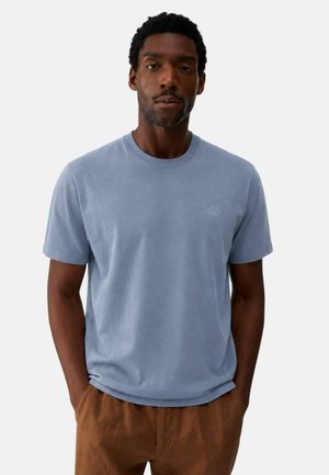 Uomo con capelli corti e ricci che indossa una T-shirt azzurro chiaro a maniche corte e pantaloni marroni, in piedi con le mani nelle tasche su sfondo bianco.